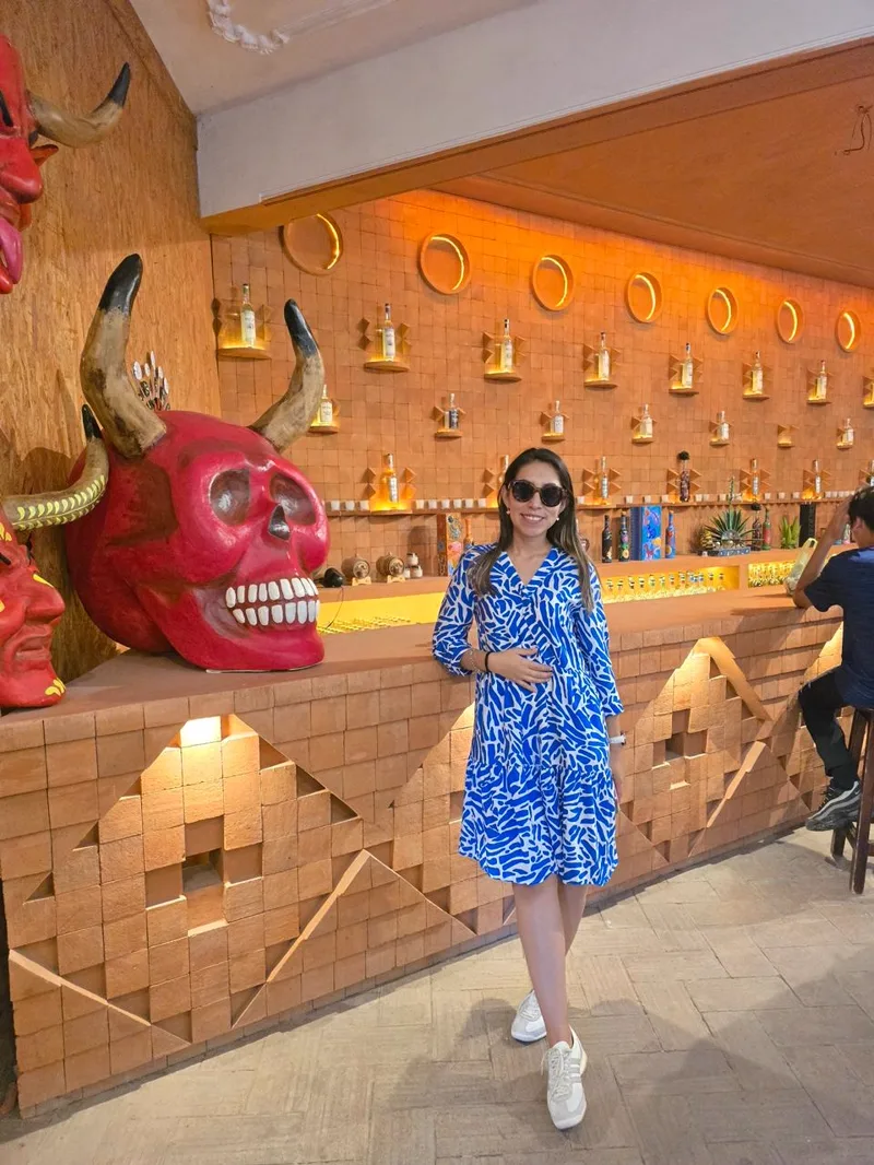 Sandy de pie en una mezcalería, junto a una calavera de cartonería